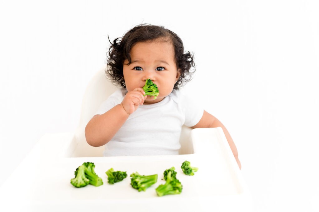 A baby happily eats broccoli in a high chair, showcasing iron-rich vegetable options for toddlers starting solids.