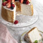 A beautifully decorated angel food cake with fresh fruit and powdered sugar sits on a crystal stand, showcasing its light and airy texture.