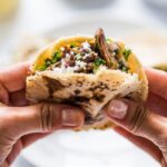 A close-up of a gordita filled with barbacoa beef, cheese, and cilantro, held in a hand.