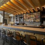 A dimly lit restaurant bar area with patrons seated at the bar and tables, showcasing a lively atmosphere.