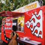 A friendly woman serves pizza slices from a food truck window, highlighting the personalized service of food trucks catering.