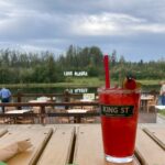 A red cocktail with a straw and a cherry on a table on a deck next to a river, with the Love Alaska Sign in the background