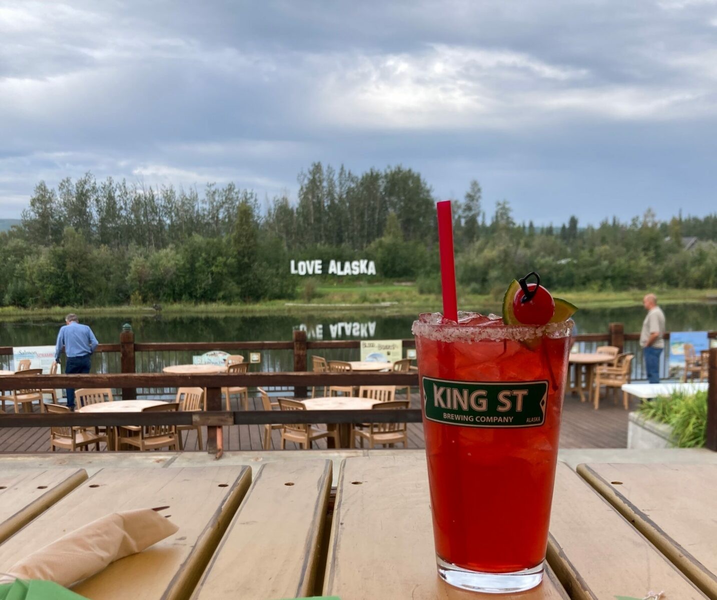 A red cocktail with a straw and a cherry on a table on a deck next to a river, with the Love Alaska Sign in the background