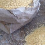 A small brown bag with sides folded down and filled wil amaranth grain. Its next to a small mound of amaranth grain