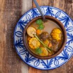 A steaming bowl of Sancocho, a traditional Dominican stew with visible chunks of meat and vegetables, garnished with cilantro.