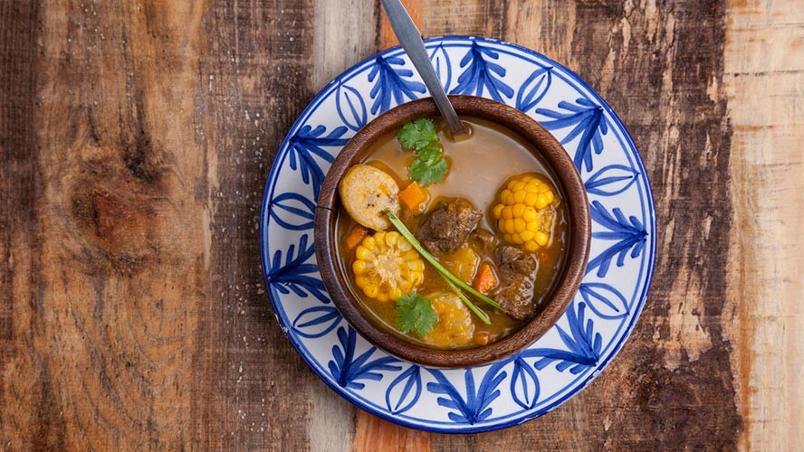 A steaming bowl of Sancocho, a traditional Dominican stew with visible chunks of meat and vegetables, garnished with cilantro.