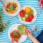 A vibrant spread of picnic food including sandwiches, salads, and fruits, laid out on a checkered blanket in a sunny outdoor setting.