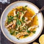 Bowl of Feel Better Chicken Soup with Lemon and Ginger, garnished with fresh herbs, in a cozy kitchen setting.