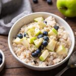 Bowl of oatmeal with blueberries and green apple chunks, alongside a small bowl of berries and a halved apple.