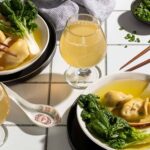 Close-up of a bowl of wonton soup with bok choy and dumplings, paired with a glass of cider