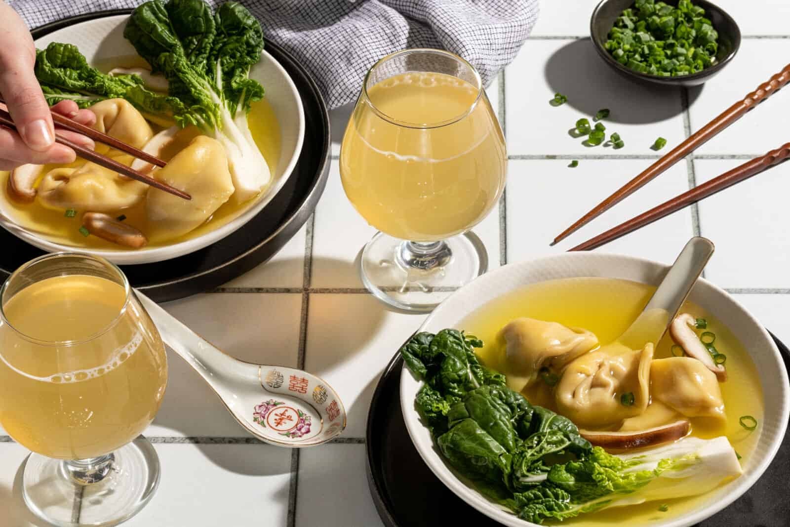 Close-up of a bowl of wonton soup with bok choy and dumplings, paired with a glass of cider