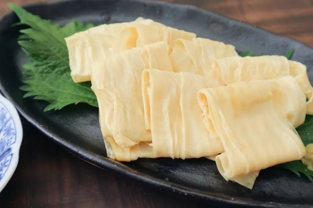 Delicate layers of yuba elegantly arranged with shiso leaves on a black plate, showcasing its refined presentation.