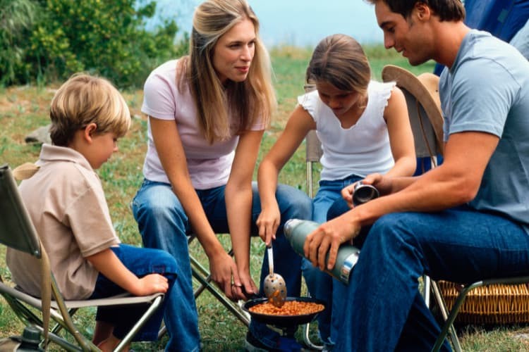 Family enjoying a camping meal of beans and sausages around a campfire