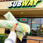 Hand Holding Two Footlong Subs in Front of a Subway Chain