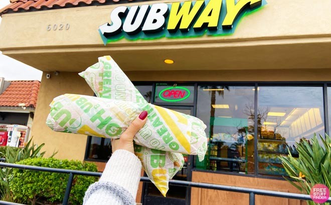 Hand Holding Two Footlong Subs in Front of a Subway Chain