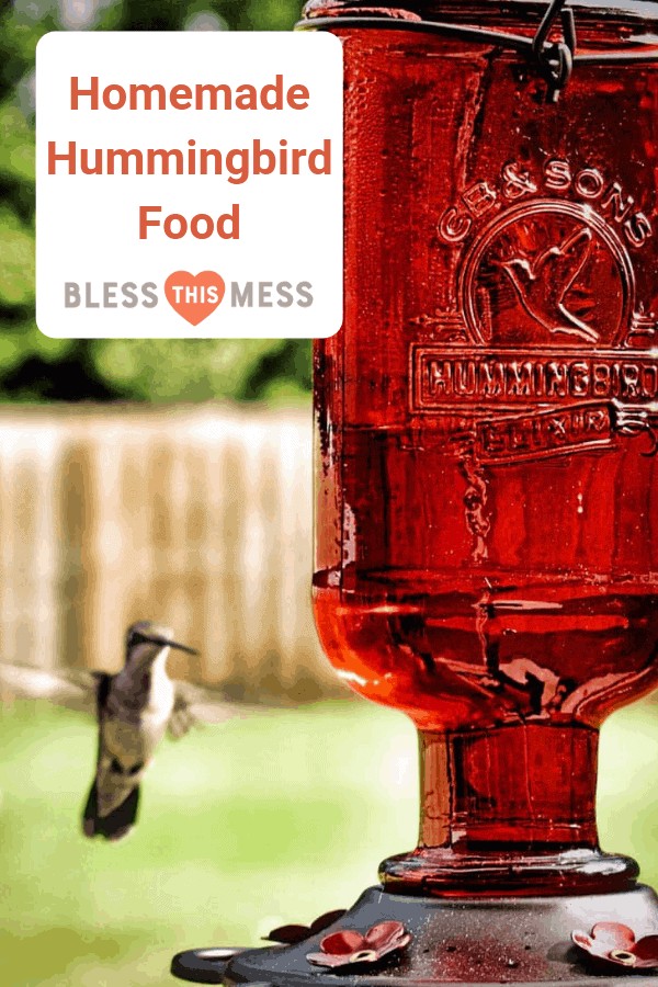 Homemade hummingbird food in a clear feeder hanging in a garden, surrounded by green foliage and colorful flowers.