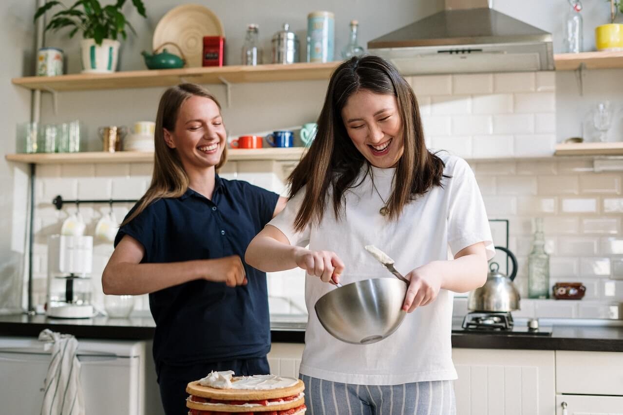 Two women laughing while making a cake, representing the enjoyment of occasional treats but also highlighting baked goods as potentially high in glucose.