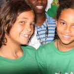 Two young girls, Sadie and Safiyah, standing outdoors in front of a vibrant green leafy backdrop, looking directly at the camera with curious and engaged expressions.