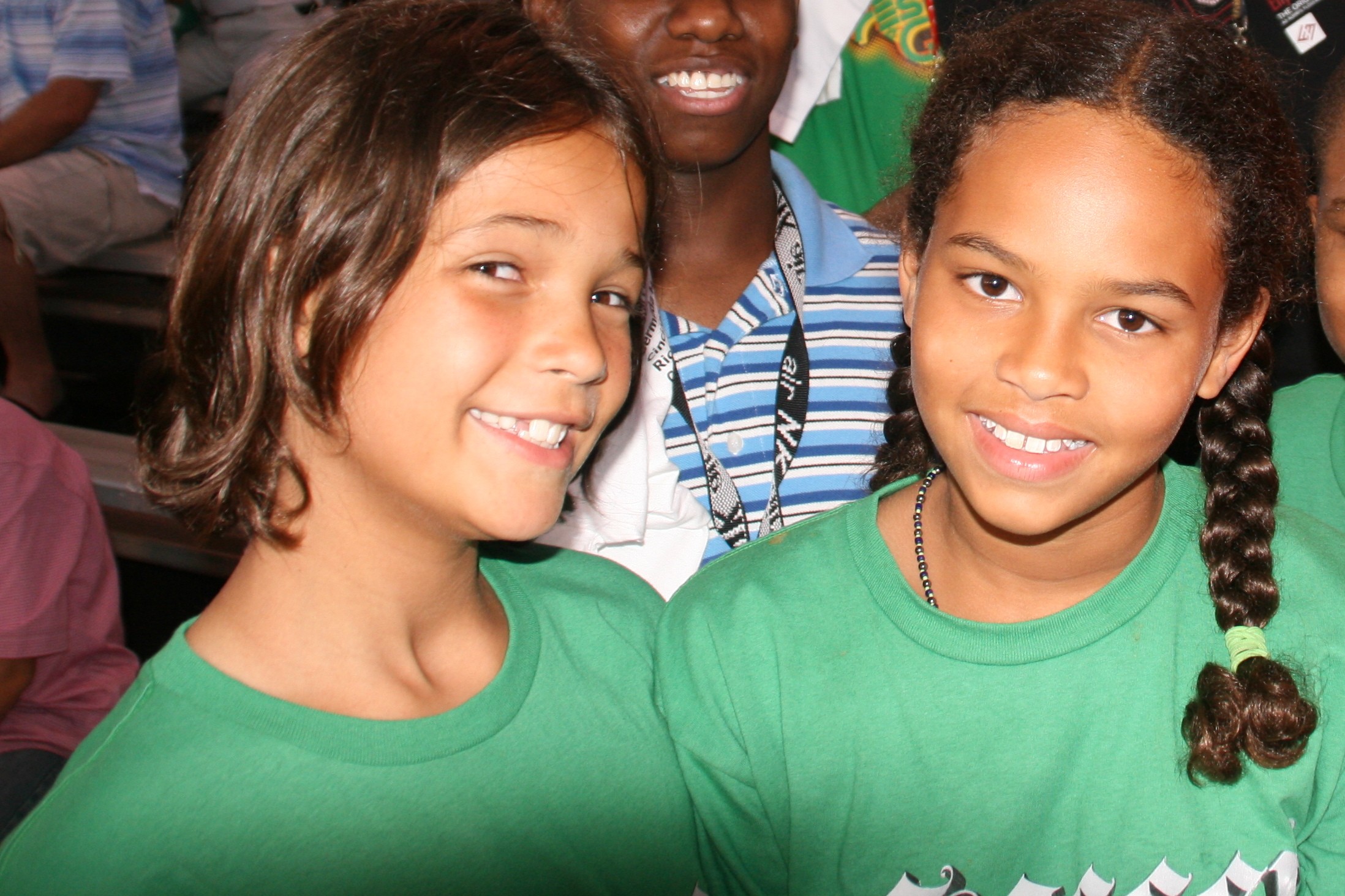 Two young girls, Sadie and Safiyah, standing outdoors in front of a vibrant green leafy backdrop, looking directly at the camera with curious and engaged expressions.