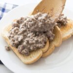 Wooden spoon scooping creamy hamburger gravy onto toasted bread slices