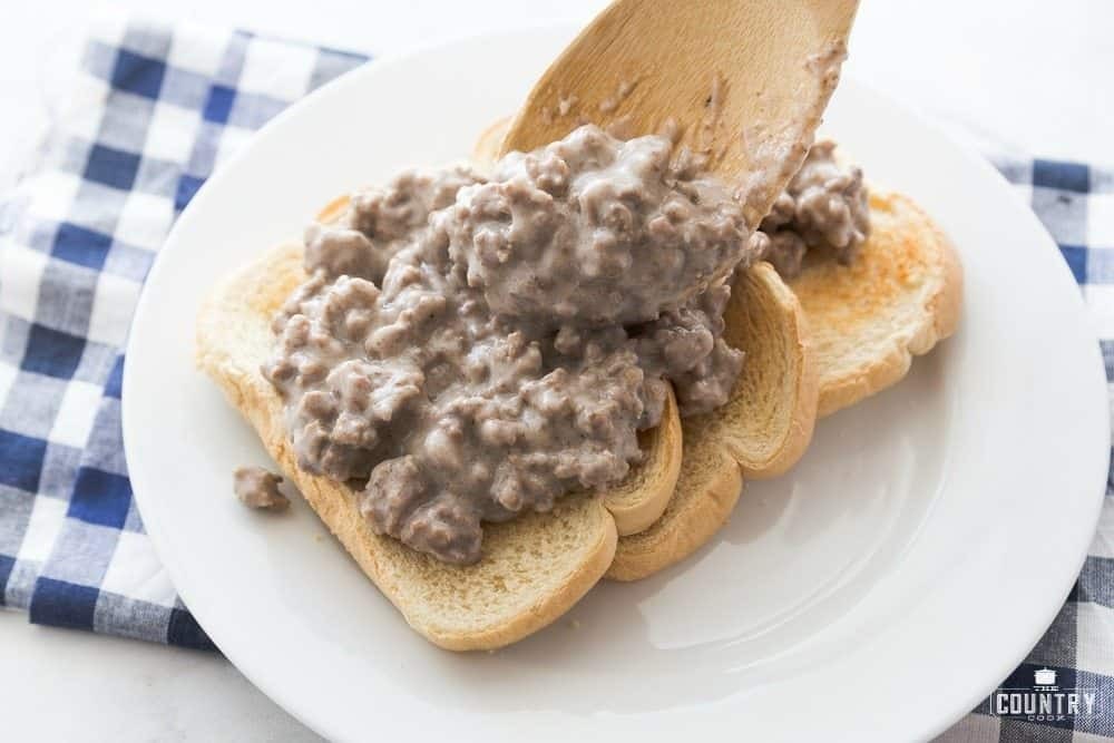 Wooden spoon scooping creamy hamburger gravy onto toasted bread slices