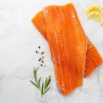photo of fresh uncooked lake trout fillets on a table top alongside lemon slices, peppercorns, and rosemary leaves