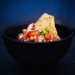 Diced shrimp, red peppers, cucumber, and red onion in a black bowl with a tostada shell for scooping.