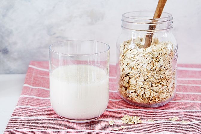 Oat milk in a glass next to a jar of oats with a spoon. A glass of oat milk sits beside a jar of oats and a spoon.