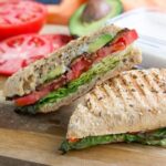 A close-up of a vegan TTLA sandwich on a cutting board, showcasing the layers of tempeh bacon, tomato, lettuce, and avocado.