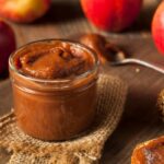 Jar of apple butter on wooden surface with apples and spoon in the background.