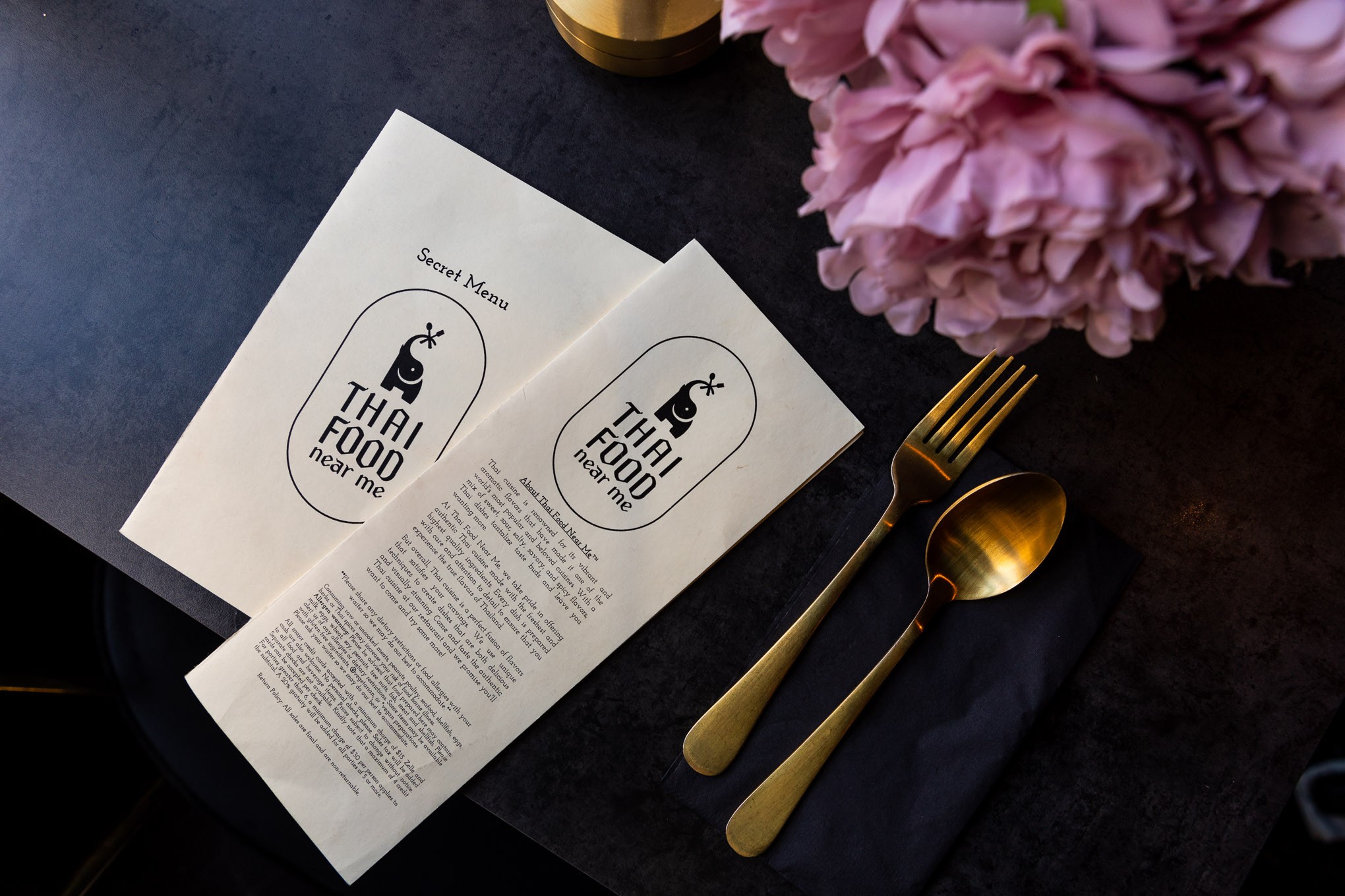 Thai Food Near Me menus are laid out on a black table with gold silverware and flowers.