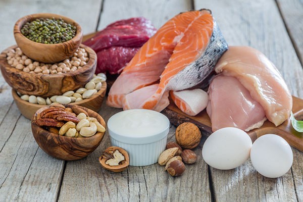 An array of protein-rich foods including beef tenderloin, grilled salmon fillet, chicken breasts, hard-boiled eggs, Greek yogurt, and various nuts and seeds