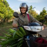 Cambodian woman smiles as she drives a motorbike loaded with green leafy vegetables