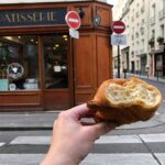 Hand holding a golden croissant in front of La Maison d'Isabelle bakery, Paris