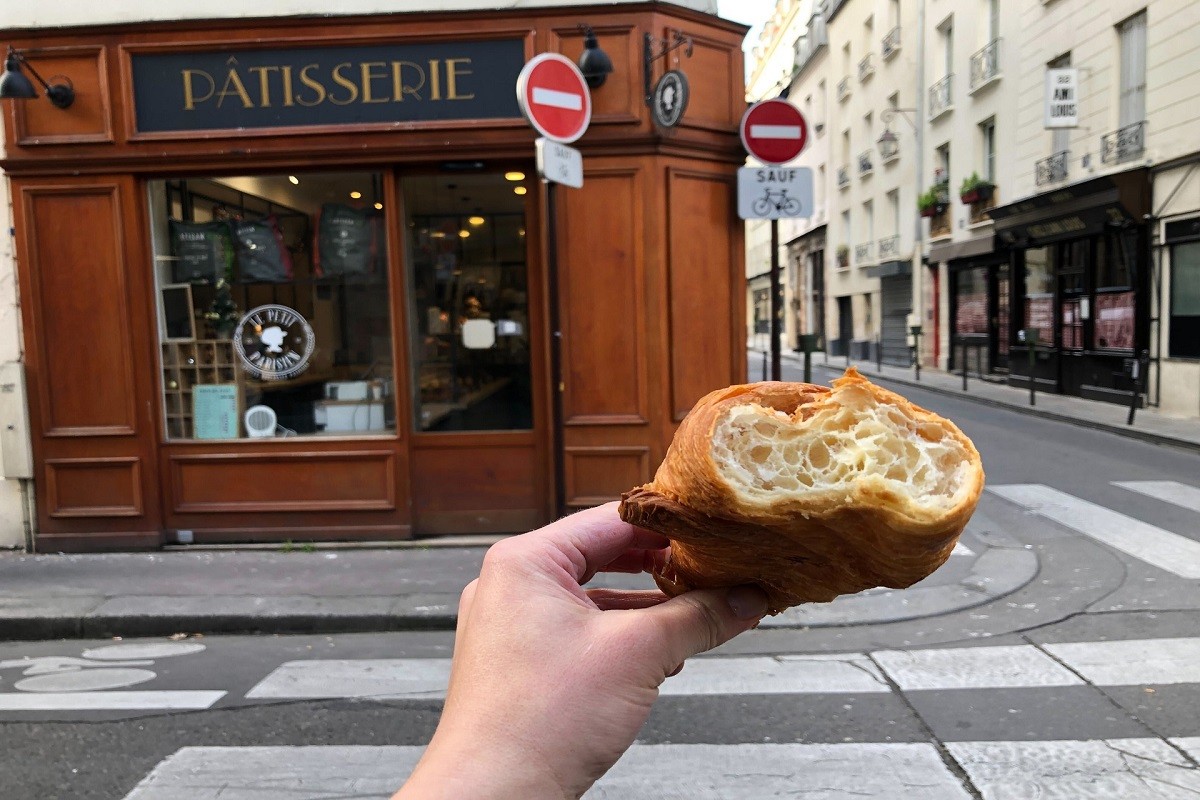 Hand holding a golden croissant in front of La Maison d'Isabelle bakery, Paris