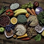 Overhead view photo of an assortment of fresh vegetables and legumes on a rustic wooden table, including corn, avocado, broccoli, orange, grapes, bell pepper, lettuce, banana, apple, almonds, and whole-grain pasta.