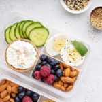 Overhead view photo of a plastic lunch box containing high-protein foods including hard-boiled egg, berries, cucumber slices, cottage cheese, and nuts