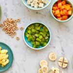 Bowls filled with finger foods arranged on a countertop, including banana slices, diced chicken, cheese cubes, and more