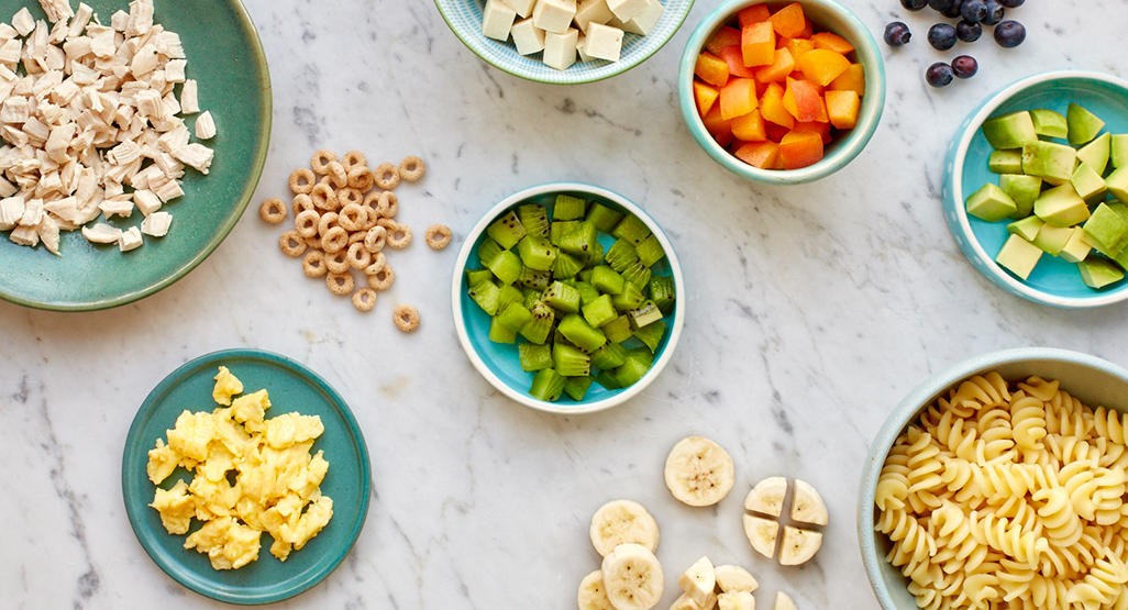 Bowls filled with finger foods arranged on a countertop, including banana slices, diced chicken, cheese cubes, and more
