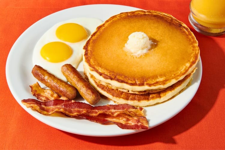 Two buttermilk pancakes served with eggs, bacon strips and sausage links.
