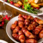 Cocktail sausages wrapped in bacon (pigs in blankets) in a white bowl on a table with other Christmas dishes partially in shot