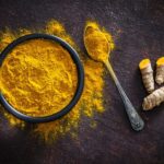 Ground turmeric powder in a bowl next to fresh turmeric root